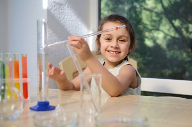Charming little girl sitting at table with laboratory glassware, holding a pipette, ready for chemical experiments in chemistry class. Measuring cylinder an test tubes with chemicals on tripod
