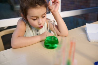 Adorable Caucasian little girl, school child watching a fascinating chemical reaction going in a flat-bottomed laboratory flask with bright green chemicals. Miracles of chemistry