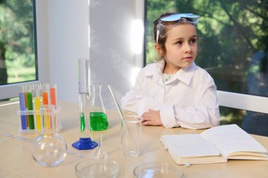 Adorable Caucasian child, little schoolgirl in protective eyeglasses and white lab coat, sitting at a table with laboratory glassware, enjoying fascinating chemical experiments in the chemistry lesson