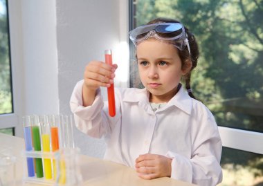 Beautiful elementary age child, smart preschool girl wearing safety goggles and white lab coat watching a chemical reaction going in the test tube she holds in her hand. Learning chemistry