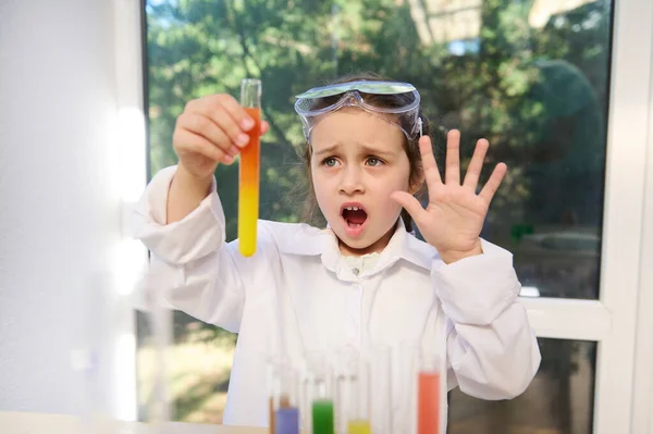 An excited little girl in protective glasses and laboratory coat, a future chemist with enthusiasm and excitement, watching a test tube with a chemical reaction going on, during the Chemistry lesson