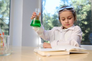 Adorable little girl has fun doing chemistry experiments, shakes a flat-bottomed flask with green solution, reads the steps of doing a science experiment. Development of children from an early age