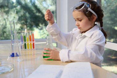 Little adorable elementary age girl using glass laboratory pipette, picking up a green solution from a flat-bottomed flask, while visiting a chemistry circle for little future chemists scientists
