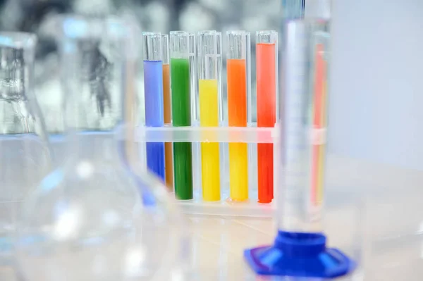 Focus on test tubes with rainbow colored chemicals and reagents standing on tripod. Blurred foreground with graduated cylinder, flasks and measuring glass labware. Chemistry, science concept
