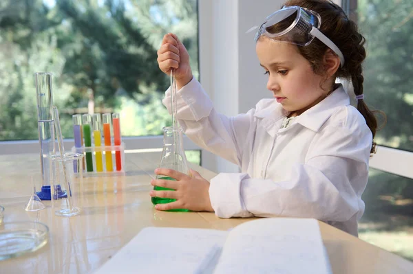 Little adorable elementary age girl using glass laboratory pipette, picking up a green solution from a flat-bottomed flask, while visiting a chemistry circle for little future chemists scientists