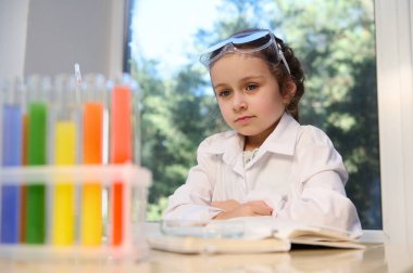 Smart schoolgirl, little chemist in safety goggles and white lab coat, sitting at the desk with textbook and test tubes with colorful chemicals on tripod, learning chemistry in the primary school