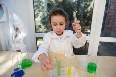 Happy inspired European preschooler girl in white lab coat pours and combines chemicals during science experiment in chemistry lesson. Graduated labware with reagents on the table in school laboratory