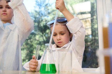 Beautiful smart little girl with pipette and test tubes making science experiment in chemistry class, standing next to a schoolboy in white lab coat in the school laboratory