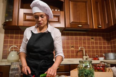 Portrait of multi-ethnic charming middle-aged woman, confident housewife in white chef cap and black apron, cooking at home kitchen, slicing raw vegetables on a chopping board. Cuisine, culinary