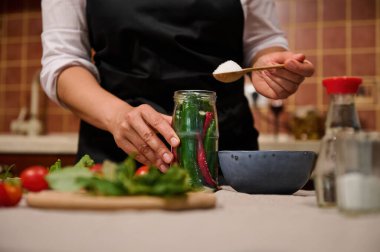 Close-up. Woman in black kitchen apron marinating hot chili, adding a scoop of white sugar for preserving it for long time, according to traditional family recipe. Fermented food. Canning. Pickling