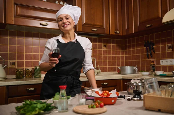 Charming middle-aged Hispanic woman, housewife in black apron and white chefs cap, sipping a glass of red wine, smiles at camera while cooking at home kitchen, preparing canned food for the winter