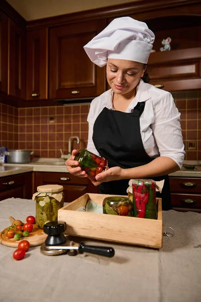 Cute woman, housewife in white chefs cap and black kitchen apron, stacking jars with homemade canned fermented and marinated vegetables in a wooden crate. Pickling according family traditional recipe