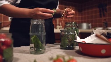 Beautiful female chef, skilled housewife in white cap and black apron filling the sterilized can for canning with fresh culinary herbs and spices, while pickling fresh organic harvest at home kitchen
