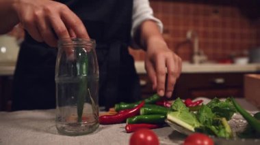 Cropped view of a housewife in a black kitchen apron, filling sterilized glass jar with spicy chili peppers and fresh chopped fragrant ingredients when pickling seasonal vegetables in the kitchen