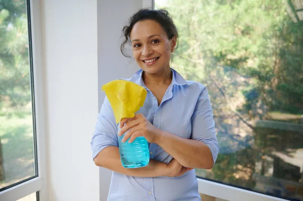 Beautiful woman of Middle-Eastern ethnicity, a pleasant housewife smiles cutely at the camera, standing against panoramic veranda windows with glass cleaner detergent and yellow rag in her hands