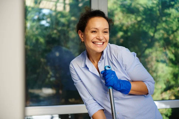 Housekeeping Job Concept. Smiling African American woman in casual blue shirt and rubber work gloves happily doing homework cleaning routine, posing with a mop and cutely smiling looking at camera