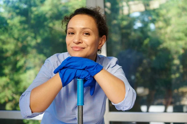 Close-up portrait of a multi-ethnic charming gorgeous happy woman, housewife in blue rubber gloves smiling at the camera, while performing a spring cleaning and keeping her house tidy. Housekeeping
