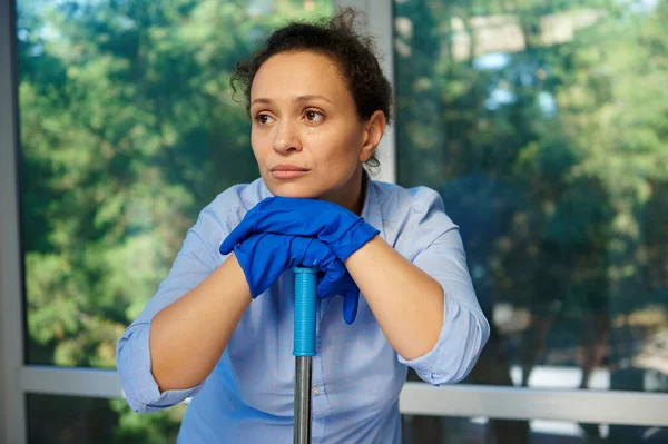 Close-up of a tired exhausted housewife, maid in blue rubber gloves, looking away thoughtfully while doing spring cleaning and keeping house in order. Cleaning service, housekeeping, domestic chores