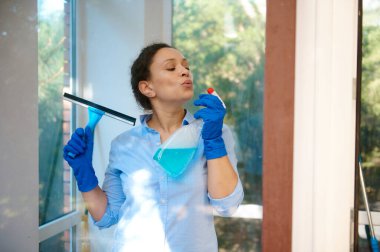 Attractive cheerful multi-ethnic woman, lovely housewife in blue rubber gloves holds glass cleaning scraper and detergent spray, happily doing household chores, washing windows, keeping her house tidy