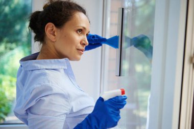 Headshot beautiful housewife with gloved hands, spraying glass cleaning household chemicals and using scraper removes stains and streaks from the windows in the veranda. Cleaning. Housekeeping. Chores