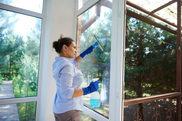 Multi-ethnic maid doing household chores wiping windows, spraying detergent and using scraper removes stains, dust and streaks while keeping house tidy. Housekeeping. Domestic life. Cleaning concept