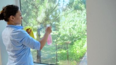 Beautiful African American woman, a lovely housewife sprinkling detergent spray and wiping panoramic window with yellow rag, happily doing household chores, keeping house tidy. Cleaning. Housekeeping