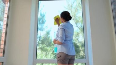Rear view. Housewife performs housekeeping chores, cleans the windows, sprays glass cleanser detergent and using a yellow rag, removes stains and streaks on the panoramic windows, keeping house tidy