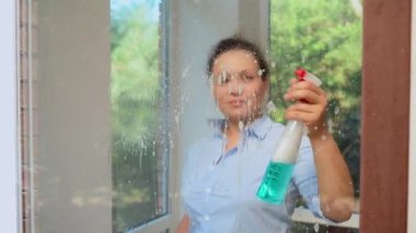 View through a glass of a pretty housewife spraying glass cleaner detergent on the windows and using a scraper to remove stains, streaks and dust during spring cleaning. Household chores. Housekeeping