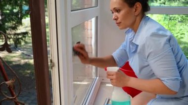 Close-up. Side view of a maid spraying glass cleaner on the window, wiping it with a rag and using a scraper to remove stains and streaks, maintaining order on the porch of the house. Household chores