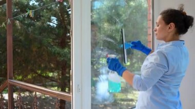 Beautiful multi-ethnic woman, a lovely housewife in blue rubber gloves, removing stains and dirt from panoramic windows on the veranda of the house. General cleaning in early spring. Household chores