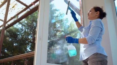 Bottom view of a charming housewife woman cleaning the panoramic windows on the veranda with a glass scraper and a spray of household chemical detergent. Maid keeping house tidy. Household chores.