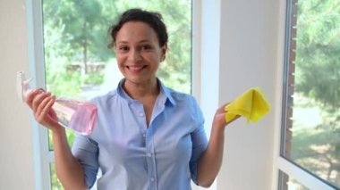 Pretty woman, housewife looking at the rag and spray detergent in her hands and spraying at the camera as at the window, wiping cleaning it. Funny housework. Emotional portrait of a lovely maid