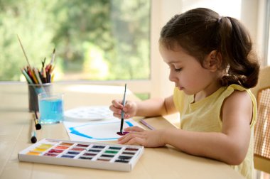 Side portrait of an adorable Caucasian little girl drawing picture while learning watercolor peinture. Talented child is panting, sitting at a table with a colorful palette and paintbrushes. Art class
