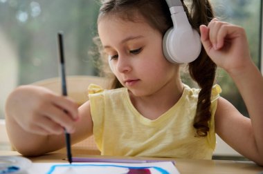 Close-up portrait of Caucasian little girl, adorable preschooler child listening to the music in wireless headphones, holding a paintbrush and painting picture with watercolors in a creative art class clipart
