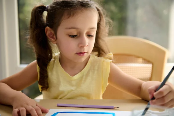 Close-up portrait of an elementary age smart little girl using a paintbrush, painting pictures with watercolor paints during an art class at home. Kids education and entertainment. Hobby and leisure