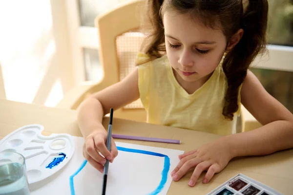 Close-up portrait of a primary school student, European smart little girl using paintbrush, painting picture with watercolor paints during art class. Kids education, entertainment, hobby and leisure