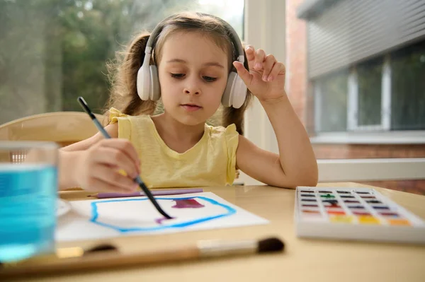 Adorable little girl wearing wireless headphones and a yellow t-shirt, listens to the music and draws picture while learning watercolor painting. Art class. Kids entertainment, hobby and leisure