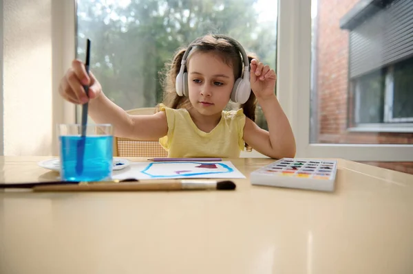 Adorable little girl wearing wireless headphones and a yellow t-shirt, listening to the music, washing a brush in a glass of water while learning watercolor painting. Art class. Hobby and leisure