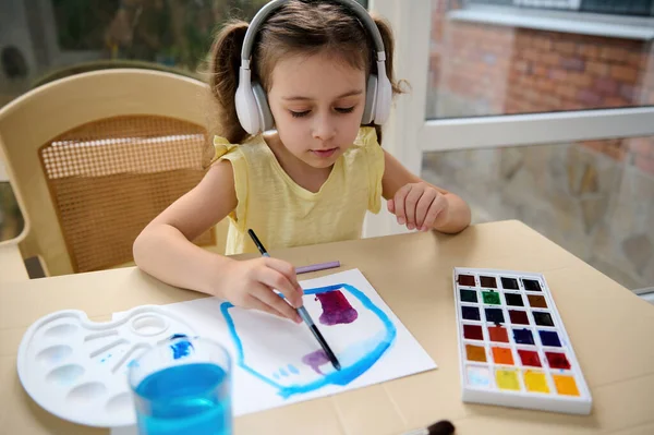 Adorable Caucasian preschooler girl in wireless headphones, concentrated little artist, drawing picture, using watercolor paints. Kids entertainment, hobby and education concept. Art master class.