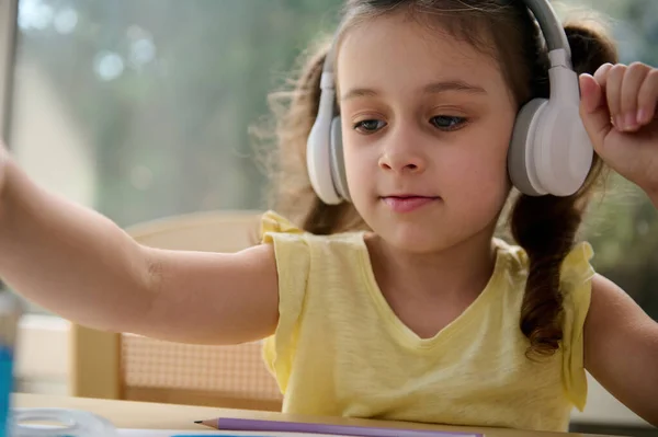 Close-up portrait of European child girl of preschool, listening to the music in wireless headphones, holding a paintbrush and painting picture with watercolors in a creative art class. Kids hobby