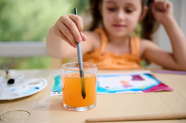 Selective focus on a glass with colored water and hand of blurred Caucasian child, preschooler girl dipping a paintbrush while drawing and painting with watercolors. Creativity. Creative art courses