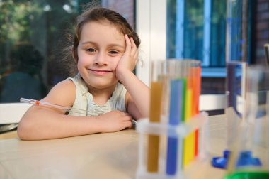 Cute preschool girl sits at a table with test tubes and glassware for a chemical laboratory, smiles sweetly at the camera. Chemistry lesson. Fascinating science experiments to keep kids entertained