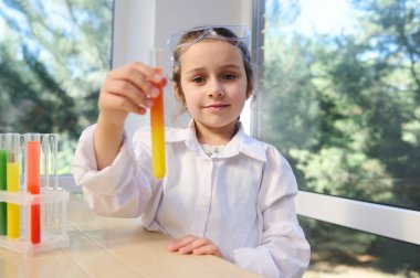 Beautiful schoolgirl of elementary school, wearing white lab coat and safety goggles, showing to the camera a test tube with going chemical reaction, at the chemistry lesson. Little scientist chemist