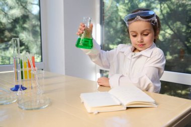 Inspired little girl, school kid wearing white lab coat and safety goggles, testing chemistry lab experiment and shaking glass tube flask, looking at chemistry textbook. Science and education concept