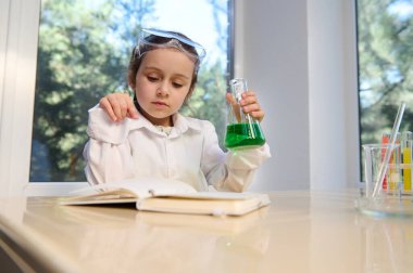 Thoughtful Caucasian little girl in safety goggles and white lab coat, concluding science experiments in chemistry class, reading text and holding a flat bottomed flask with green chemical solution