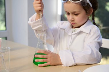 Adorable preschooler girl wearing white lab coat, enjoying learns chemistry at home laboratory, using flask and pipette, makes experiment . Fascinating scientific experiments for kids entertainment