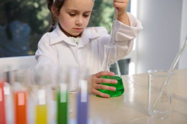 Selective focus on flat-bottomed flask with green liquid and little girls hand holding pipette, collecting some chemicals, doing science experiments in school lab during chemistry lesson