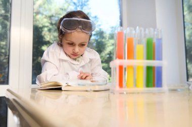 European smart preschooler girl in protective eyewear and white lab coat, future scientist chemist learns chemistry, sits at a table with test tubes with rainbow color chemical solutions on a tripod