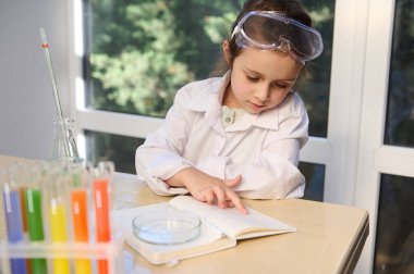 European smart little girl, future chemist in safety goggles and white lab coat, studying chemistry in the school laboratory. Fascinating science and chemistry experiments to keep kids entertained.