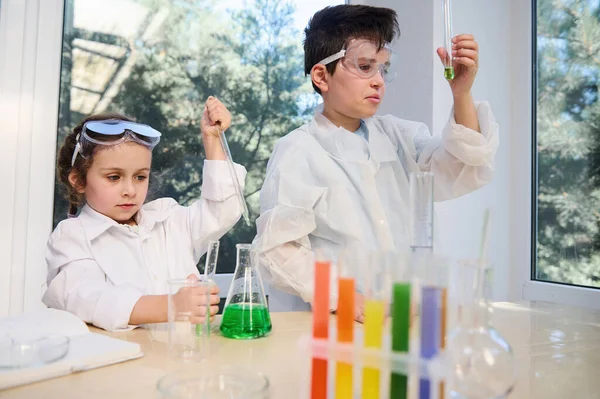 Diverse kids, teenage boy and preschooler girl in white laboratory coats and protective eyeglasses, doing biochemistry research in chemistry class. Test tubes, flasks with chemicals on the foreground